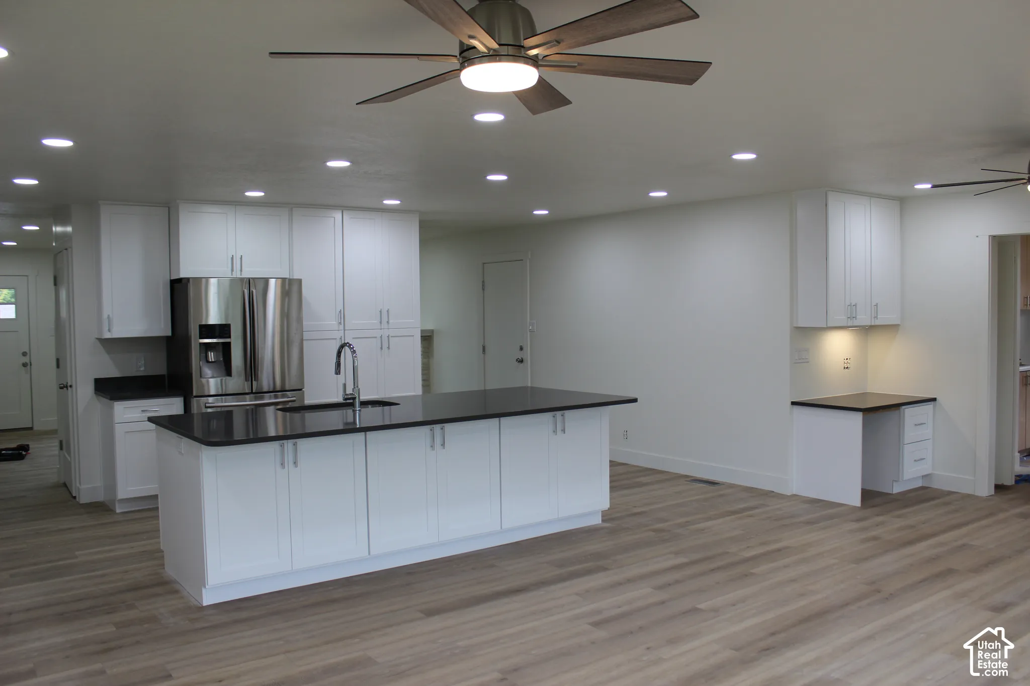 Kitchen featuring white shaker-style cabinetry, dark stone countertops, a central island with an integrated sink, recessed lighting, and wood-finish flooring