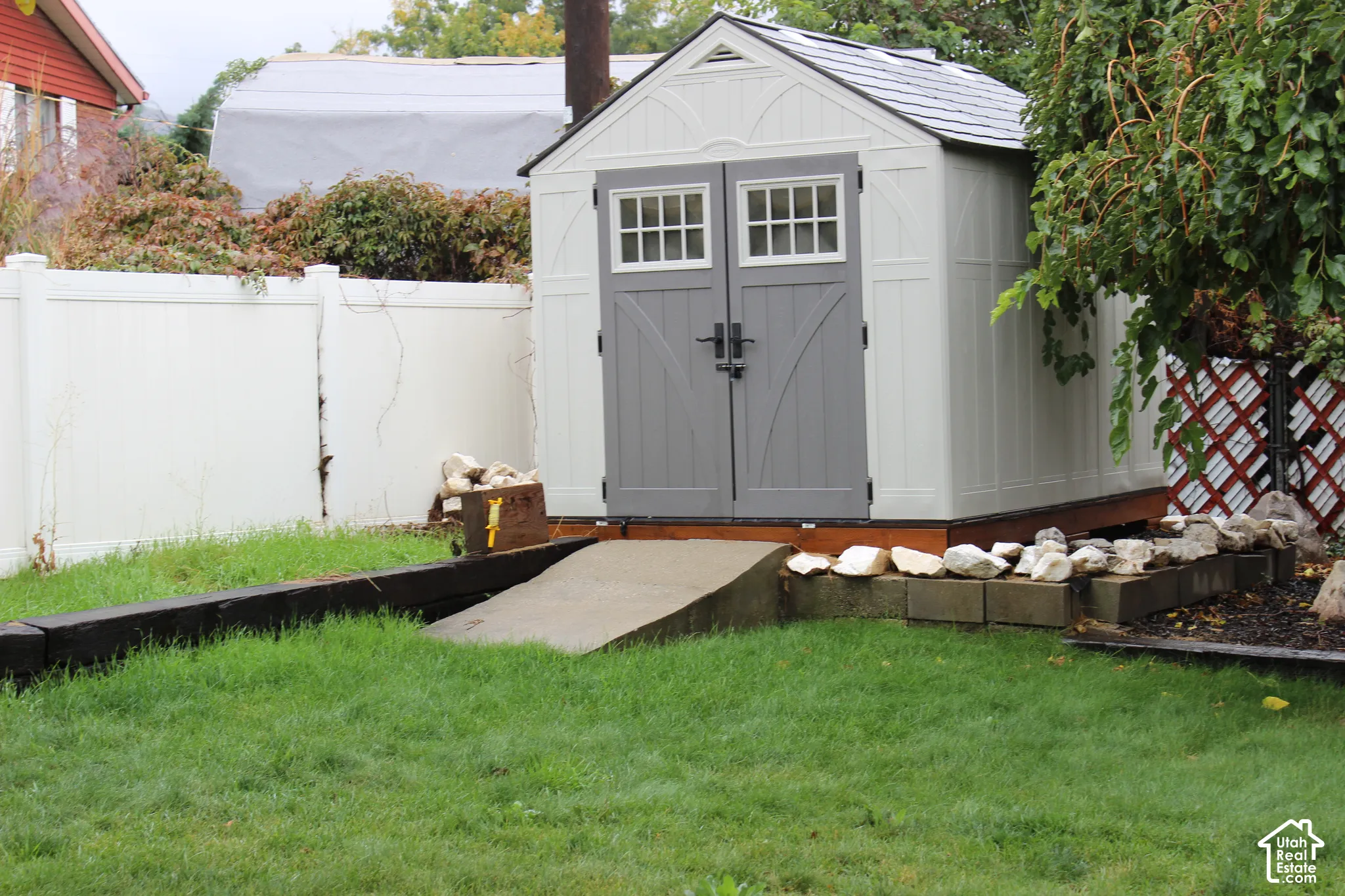 Storage shed featuring double-door entry with windows, composite exterior, and a dedicated concrete access ramp, enclosed by perimeter vinyl fencing