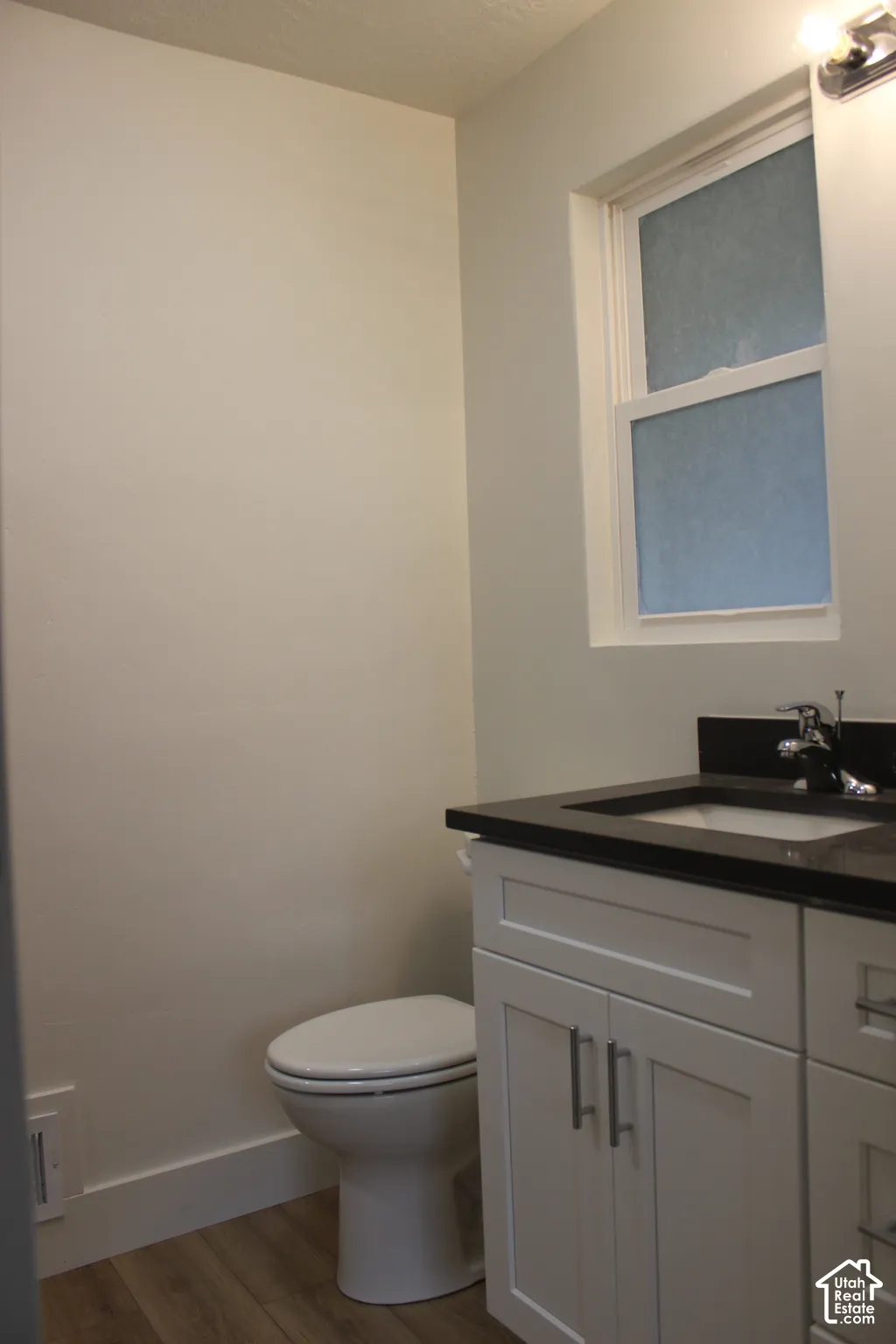 Bathroom featuring a white shaker-style vanity cabinet, dark countertop with an integrated basin, chrome faucet, and wood-finish flooring