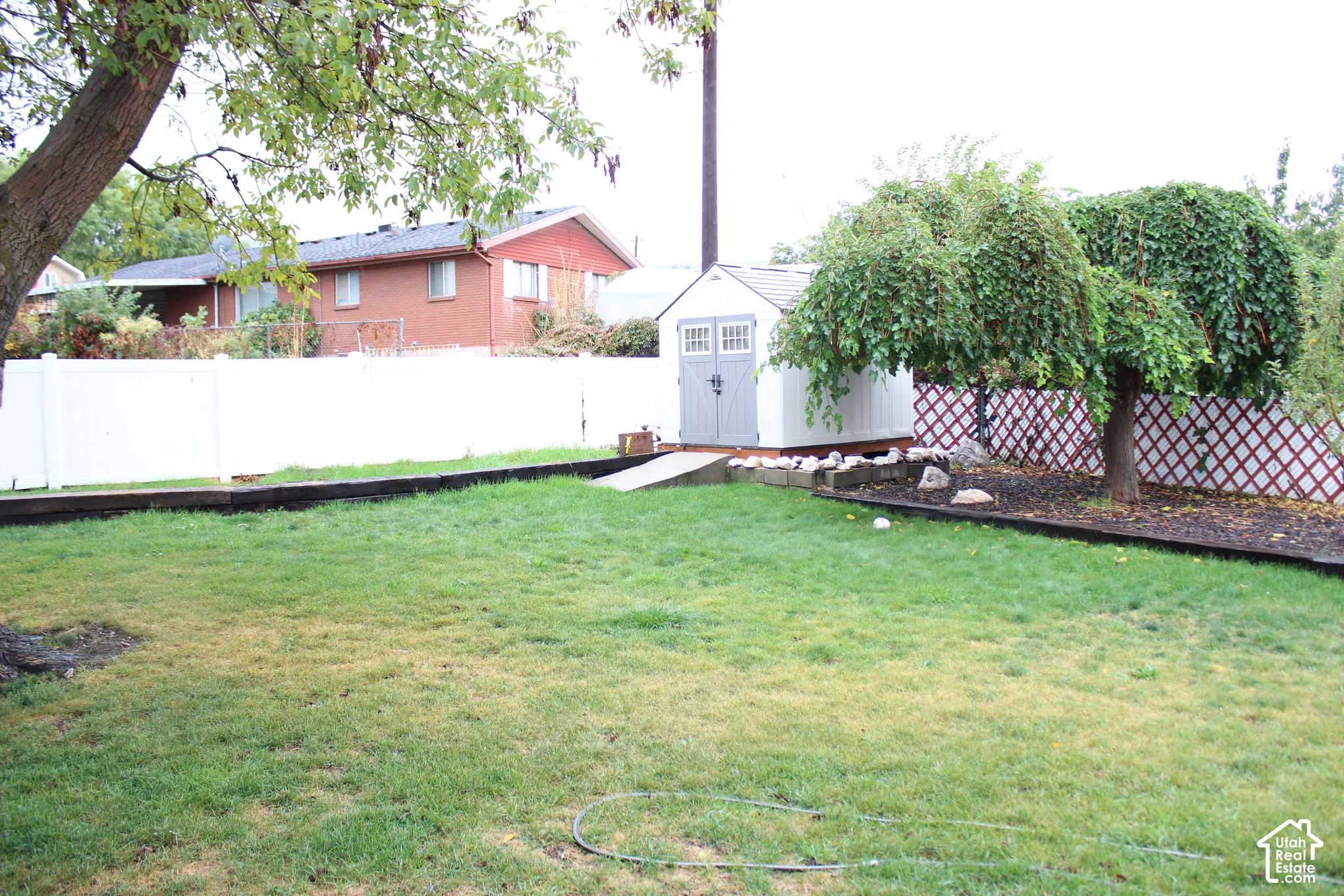 Backyard featuring a grass lawn, perimeter white vinyl fencing, storage shed, timber retaining walls, and mature landscaping