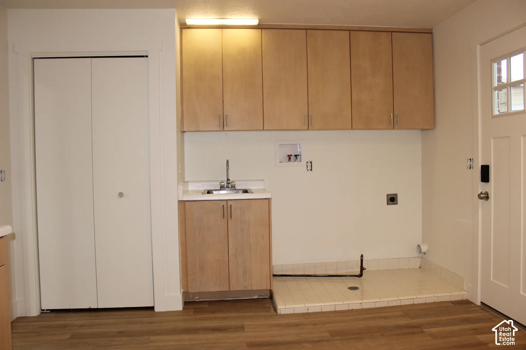 Laundry area featuring wood-finish cabinetry, a built-in sink with metal faucet, tiled utility floor, bi-fold closet doors, and wood-finish flooring