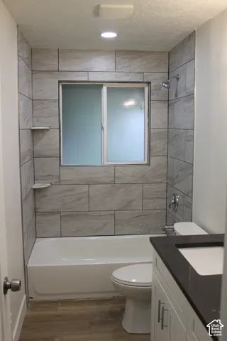 Bathroom featuring a tub and shower combination with full-height stone-finish tile surround, integrated shelving, white shaker-style cabinetry, and a dark solid-surface countertop