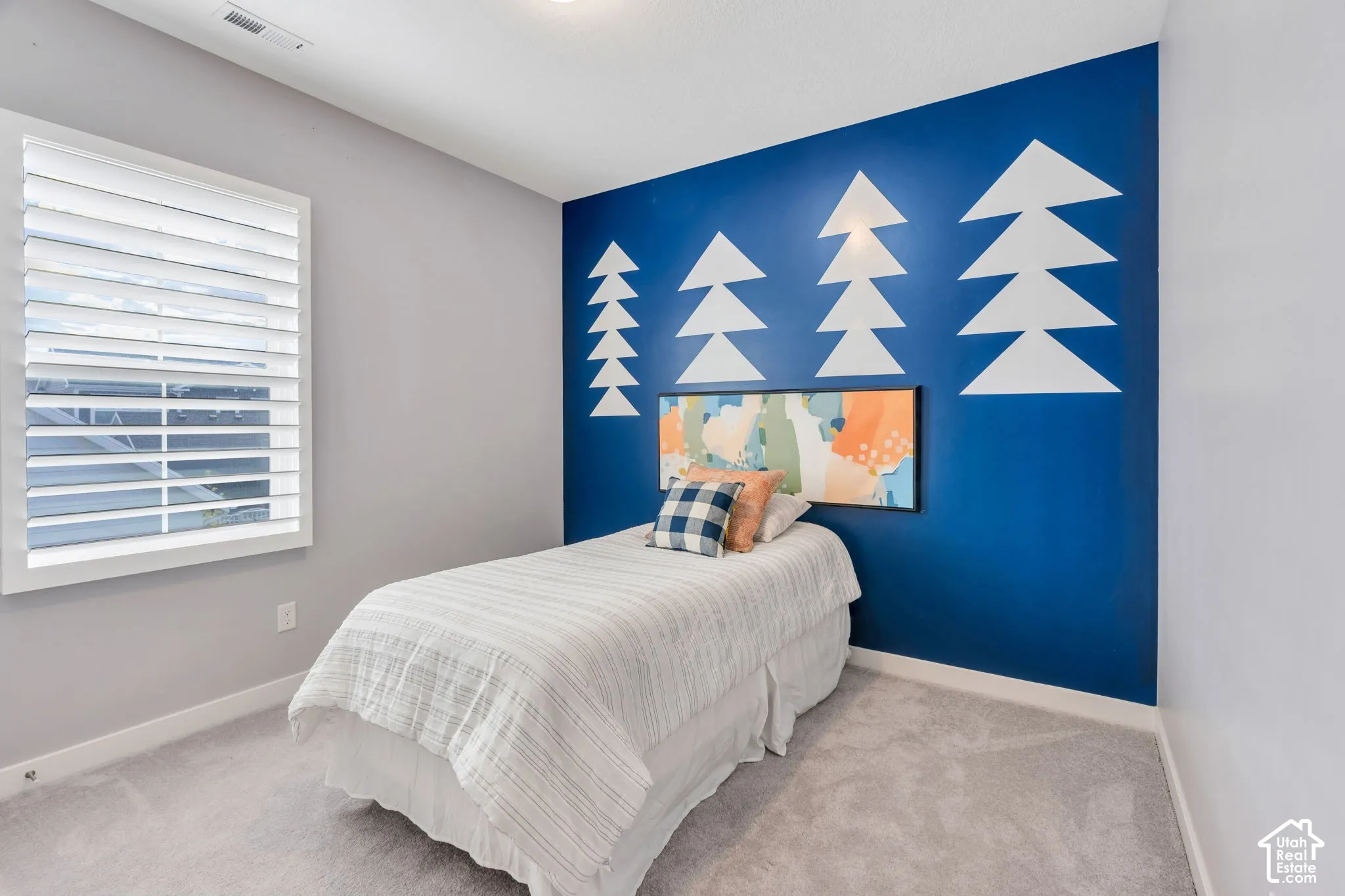Neutral-toned carpeting, accent wall with geometric painted detailing, and window fitted with horizontal plantation-style shutters