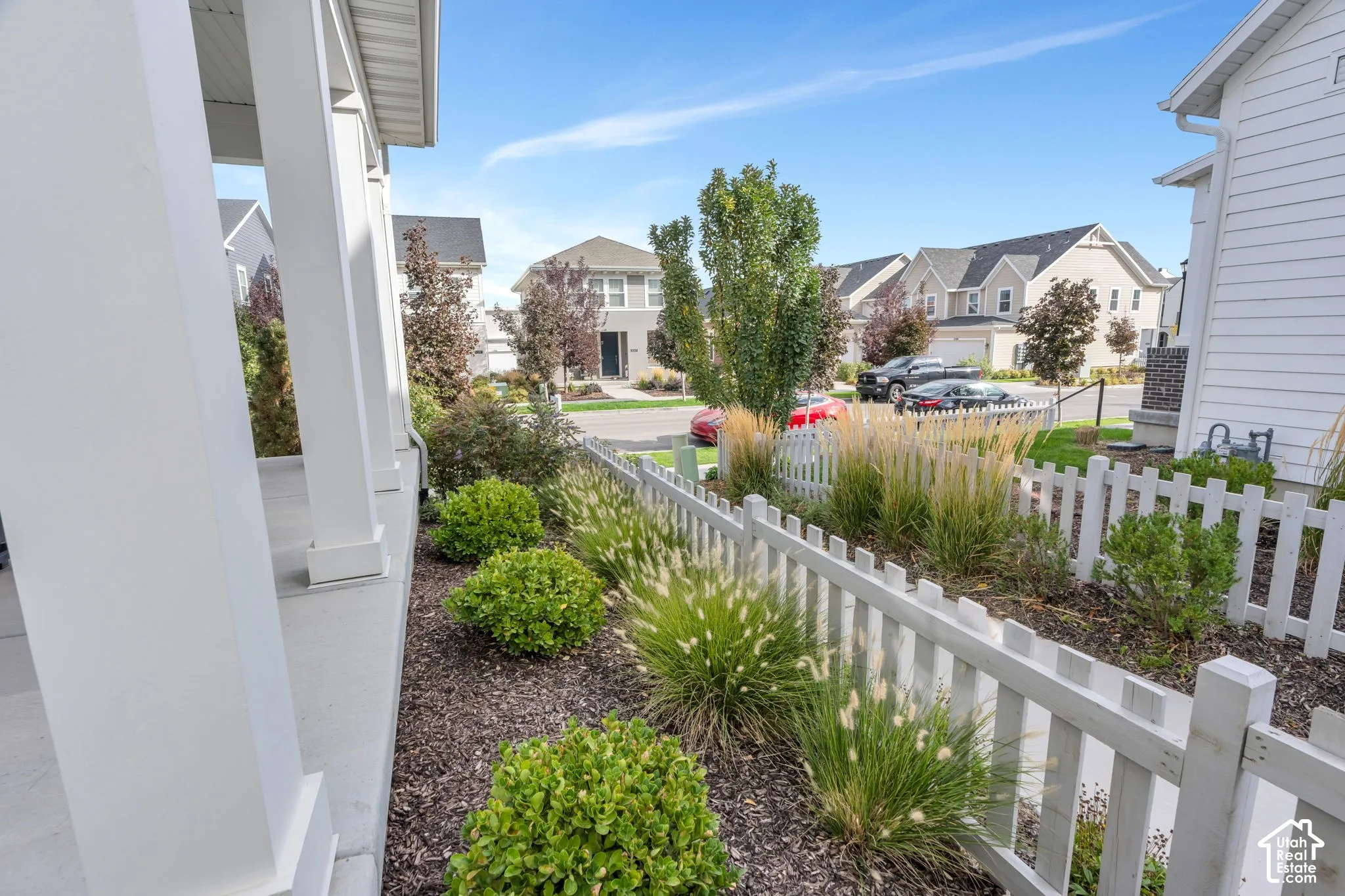 Covered front porch with structural columns, white picket fence, and landscaped garden beds featuring ornamental grasses and shrubs