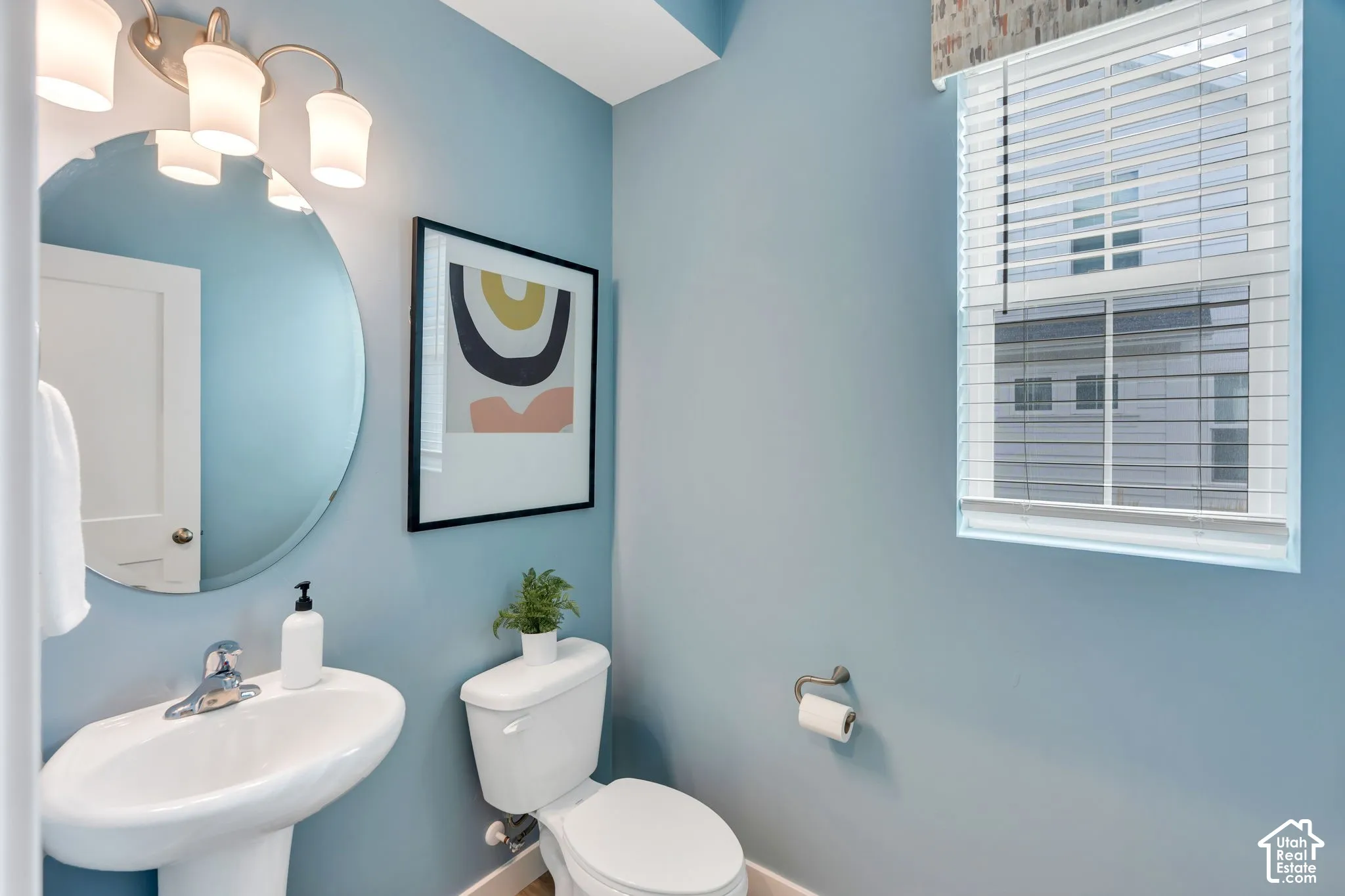 Oval wall-mounted mirror, pedestal sink with polished chrome faucet, multi-bulb vanity light fixture, and window with horizontal blinds