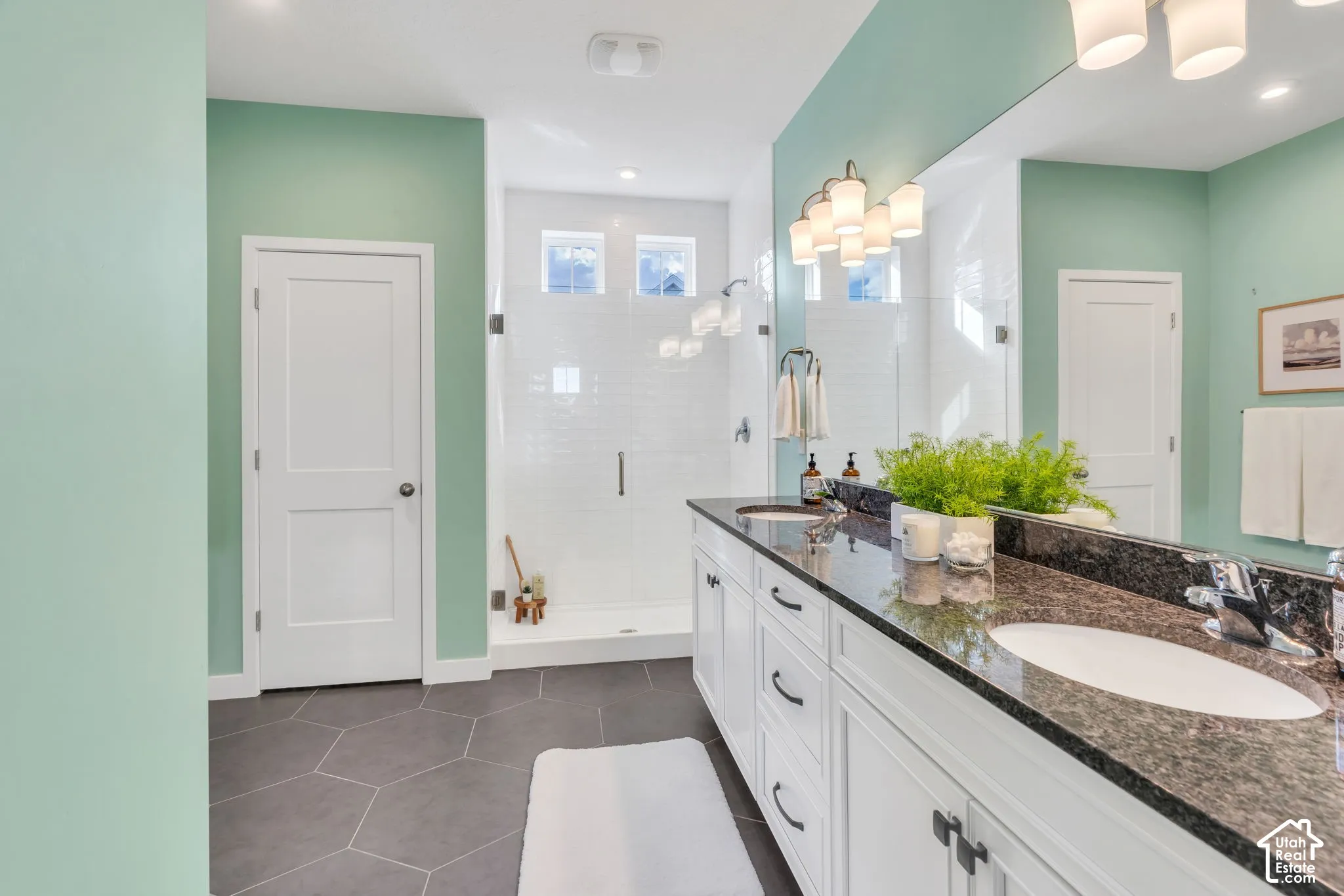 Double vanity with stone countertop and undermount sinks, walk-in shower with glass enclosure, large wall-mounted mirror, and hexagonal tile flooring
