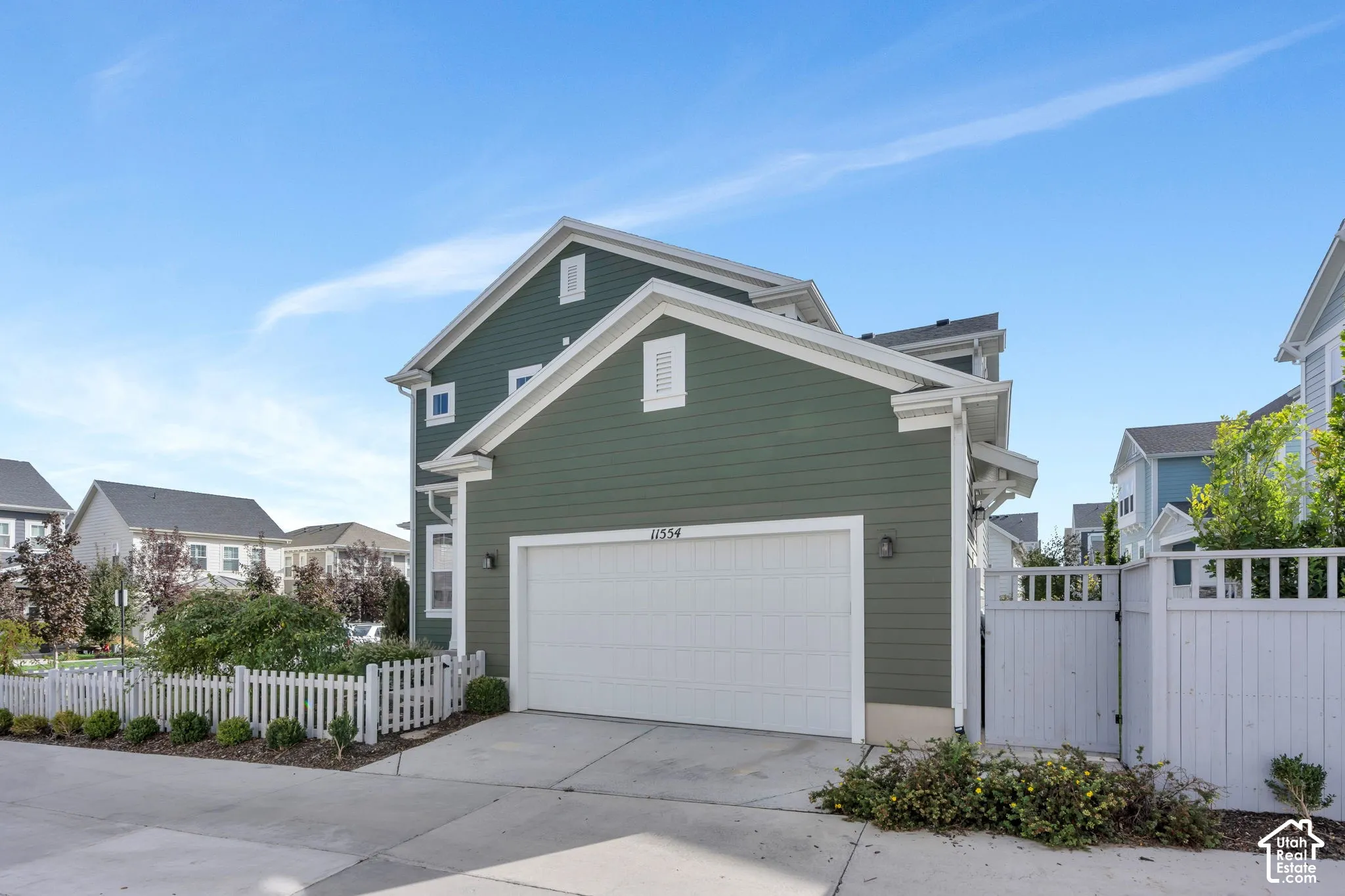 Multi-level exterior featuring dark green horizontal siding, a white sectional garage door, a white picket fence, and a private wooden gate