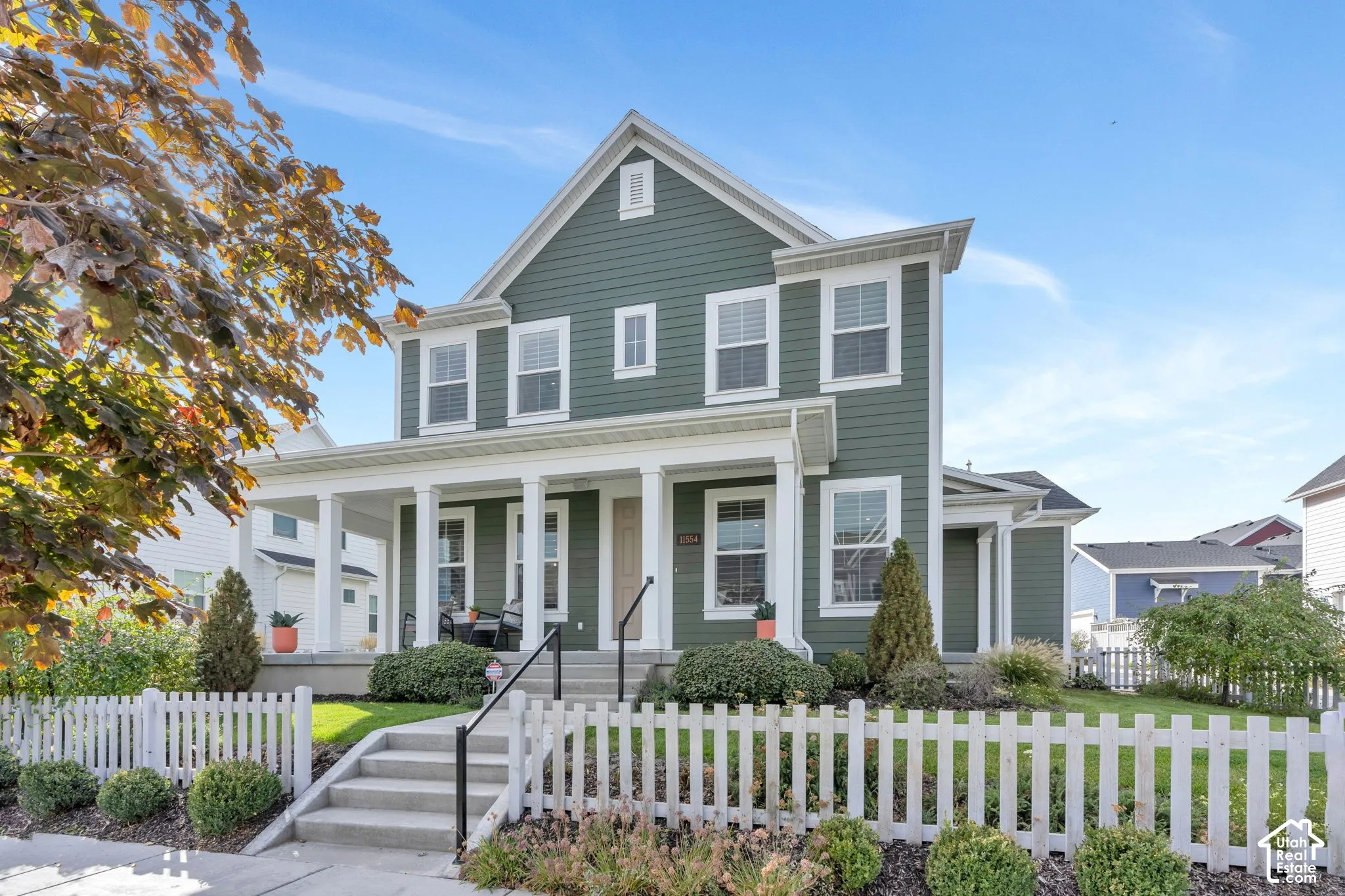 Two-story exterior featuring sage green horizontal siding, white trim, a covered front porch with classical columns, and a white picket fence enclosing the front yard