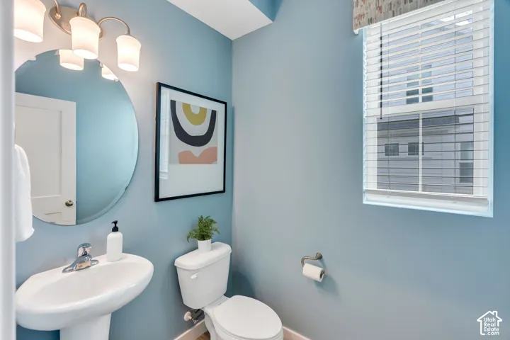 Oval wall-mounted mirror, pedestal sink with polished chrome faucet, multi-bulb vanity light fixture, and window with horizontal blinds