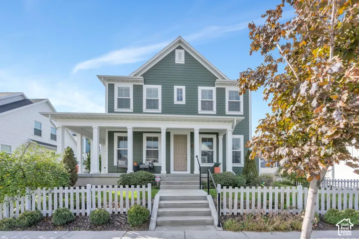 Two-story residence with sage green horizontal siding, white trim, a covered front porch with square columns, and a white picket fence enclosing the front yard
