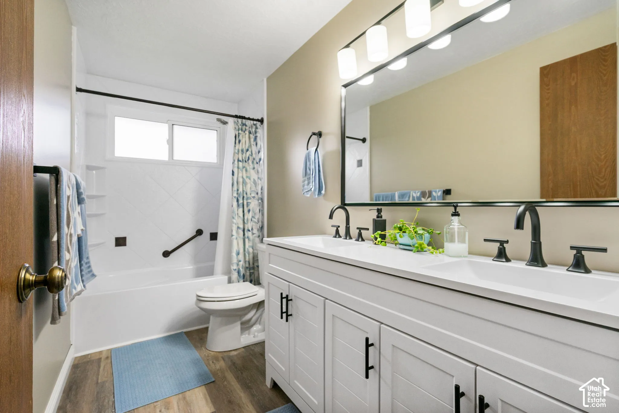 Bathroom featuring a dual-sink vanity with white cabinetry, matte black hardware, a large framed vanity mirror, integrated multi-bulb lighting, and wood-finish flooring