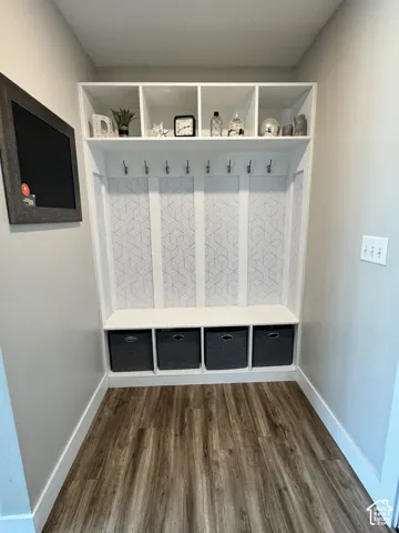 Built-in mudroom storage featuring an integrated bench, four open-front storage cubbies, eight brushed-nickel coat hooks, and decorative geometric-patterned paneling, finished with wood-finish flooring