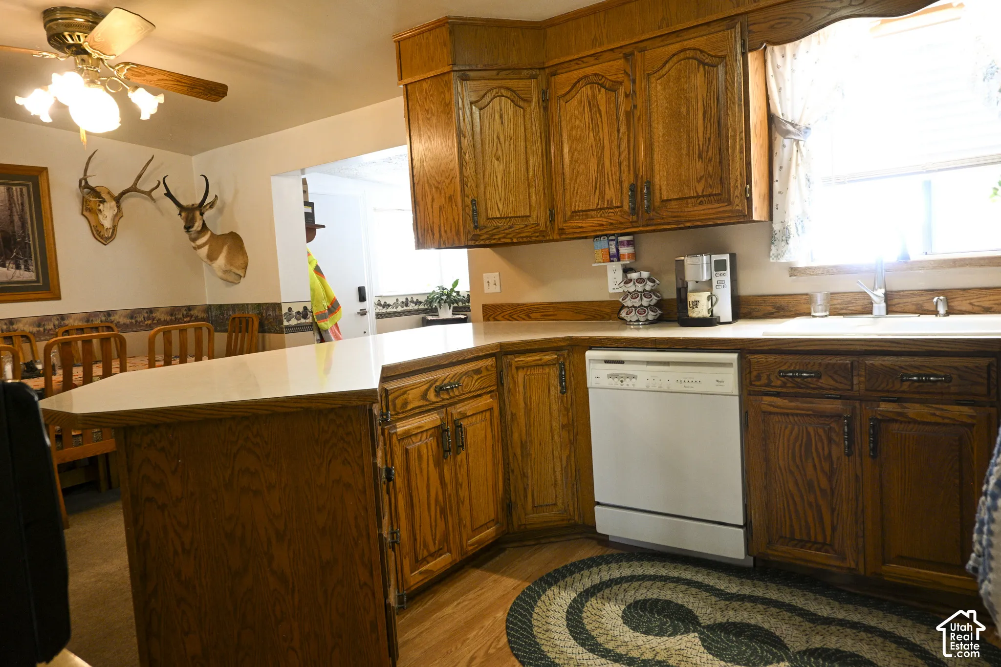 Kitchen featuring stained wood cabinetry, solid surface countertops, peninsula with counter seating, integrated white dishwasher, and ceiling-mounted light fixture with fan