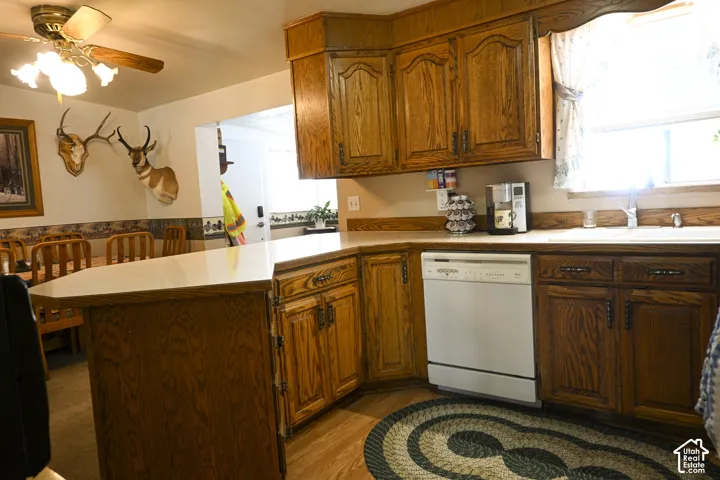 Kitchen featuring stained wood cabinetry, solid surface countertops, peninsula with counter seating, integrated white dishwasher, and ceiling-mounted light fixture with fan