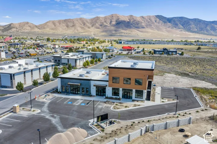 Modern mixed-use building featuring horizontal wood-cladding, dark metal facade accents, expansive glass storefronts, flat roof architecture, and an asphalt parking area with designated accessible spaces against a mountain backdrop