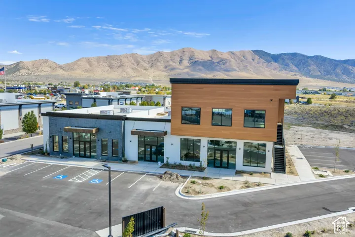 Multi-tenant commercial building featuring a modern wood-paneled upper facade, dark brick detailing, large glass storefronts, and an adjacent asphalt parking area with mountain backdrops