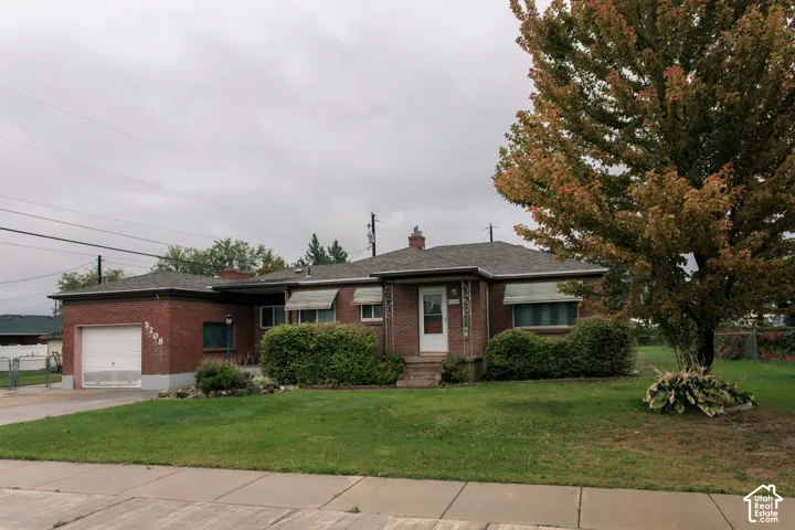Brick exterior residence featuring an attached garage, shingled roof, metal window awnings, and mature landscaping
