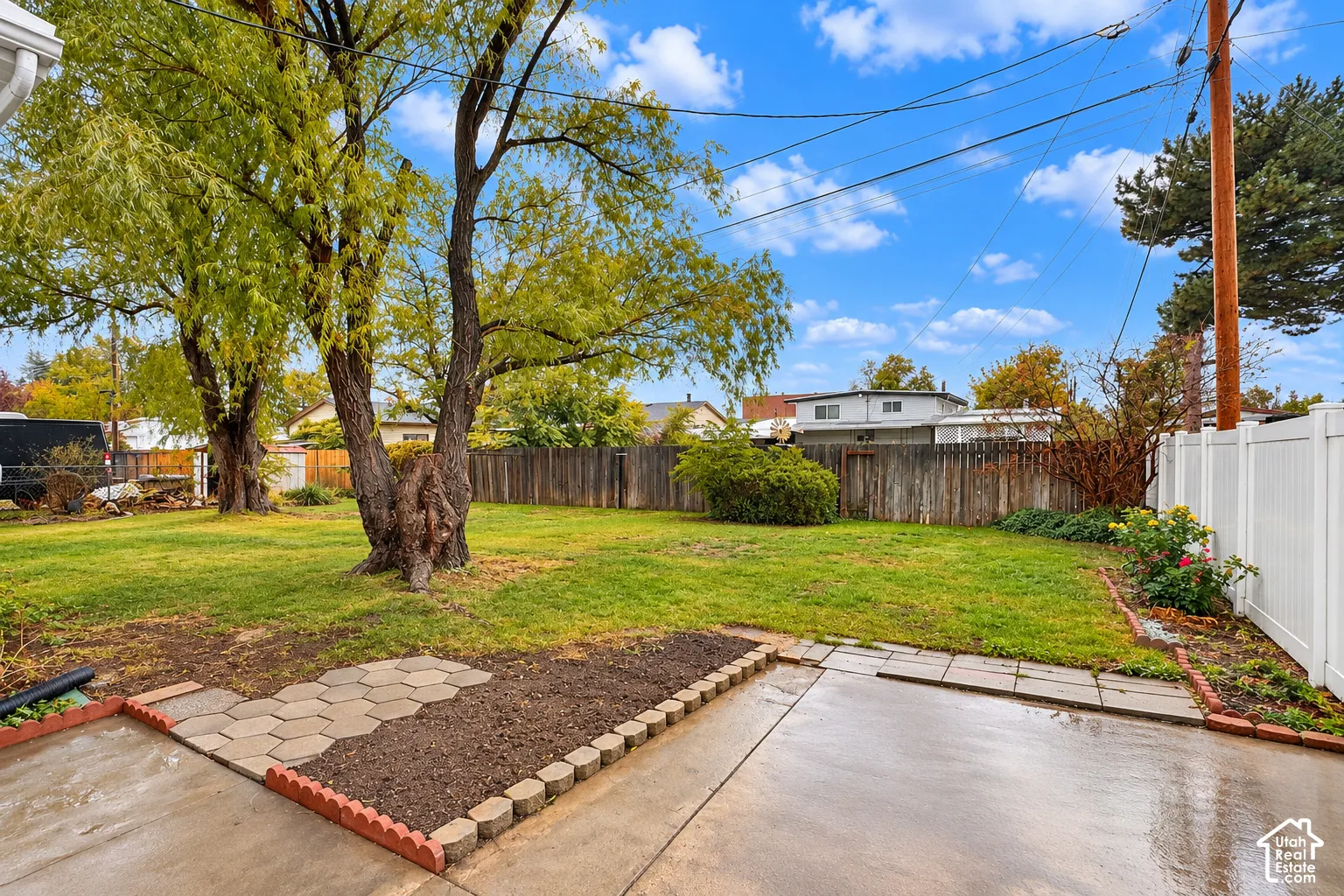 Backyard featuring a concrete patio, stone-edged planting beds, a sod lawn, mature trees, and perimeter wood and vinyl fencing