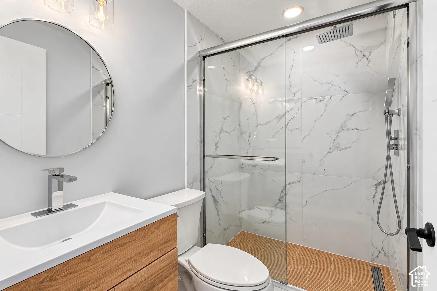 Bathroom featuring a wood-finish vanity with a white countertop, circular wall-mounted mirror, walk-in shower with floor-to-ceiling stone-style tile, rainfall showerhead, and a handheld attachment behind a glass enclosure