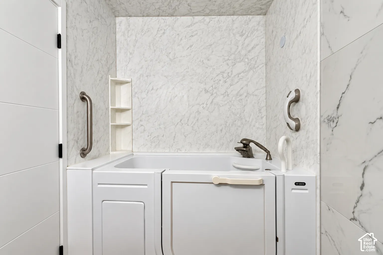 Walk-in bathtub featuring stone-patterned wall surround, metallic finish grab bars, integrated shelving, and bronze-tone faucet hardware