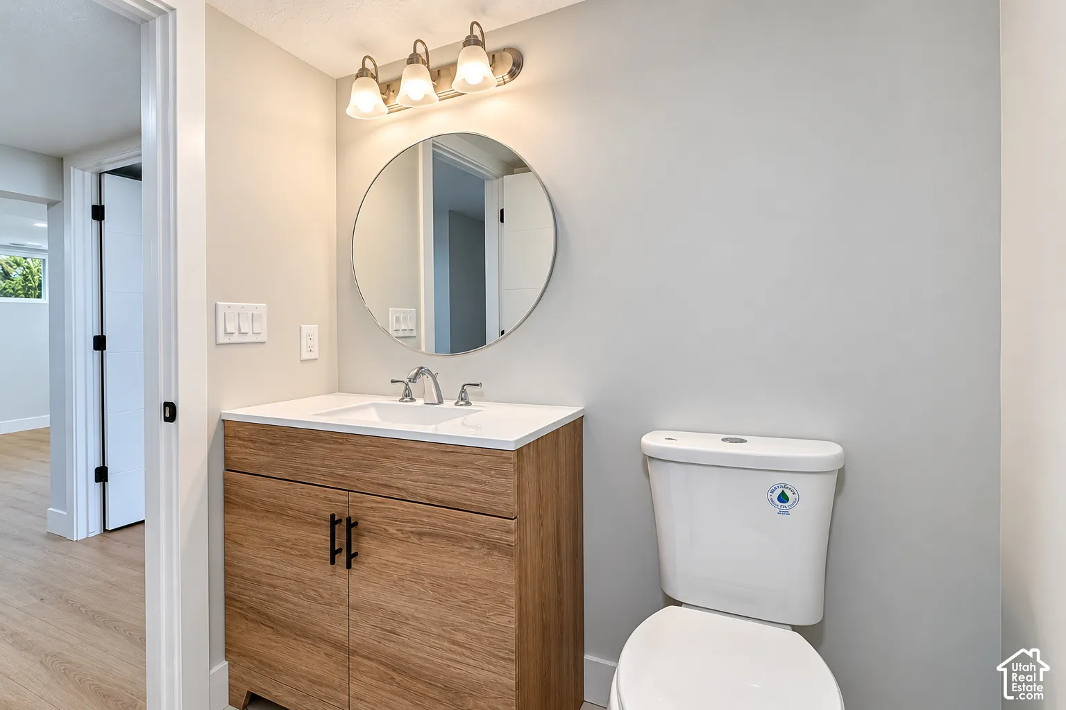 Vanity featuring a wood-finish cabinet, white solid-surface countertop, rectangular undermount sink, circular wall-mounted mirror, and a three-light fixture