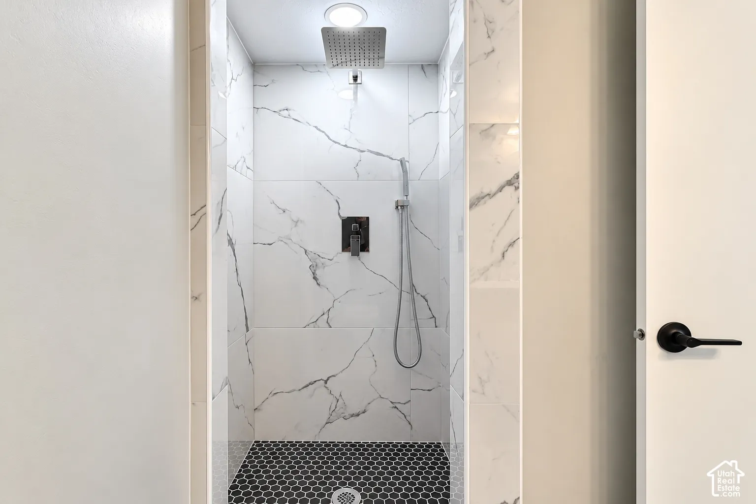 Walk-in shower featuring large-format white stone-finish wall tile with charcoal veining, black hexagonal floor tile, a rainfall showerhead, and a matte black handheld shower fixture