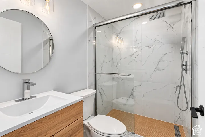 Bathroom featuring a wood-finish vanity with a white countertop, circular wall-mounted mirror, walk-in shower with floor-to-ceiling stone-style tile, rainfall showerhead, and a handheld attachment behind a glass enclosure