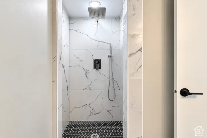 Walk-in shower featuring large-format white stone-finish wall tile with charcoal veining, black hexagonal floor tile, a rainfall showerhead, and a matte black handheld shower fixture