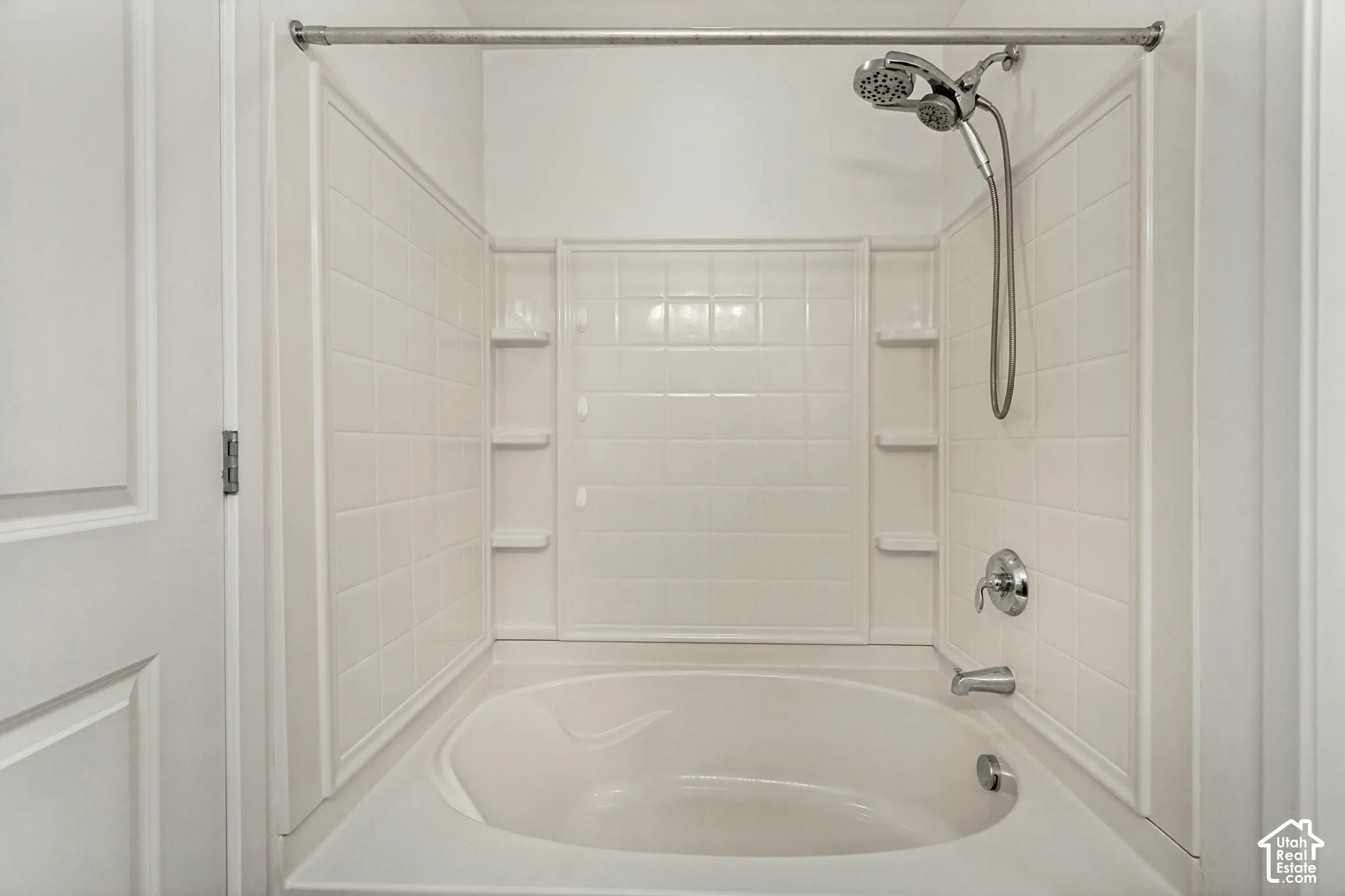 Built-in soaking tub with a tiled surround, integrated shelving, handheld shower fixture, and a metal shower rod