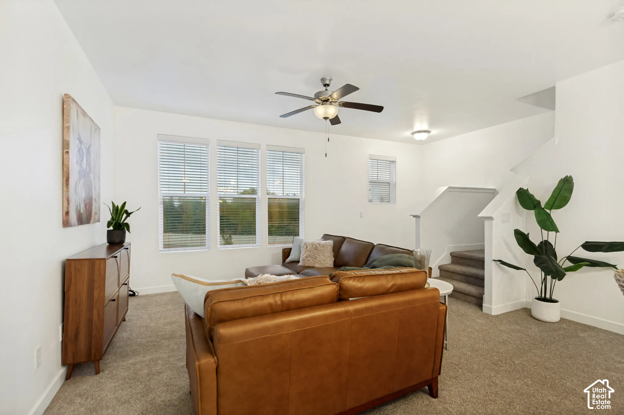 Open-concept interior featuring neutral wall tones, wall-to-wall carpeting, triple-pane window configuration with slatted blinds, and a central ceiling fan with integrated lighting