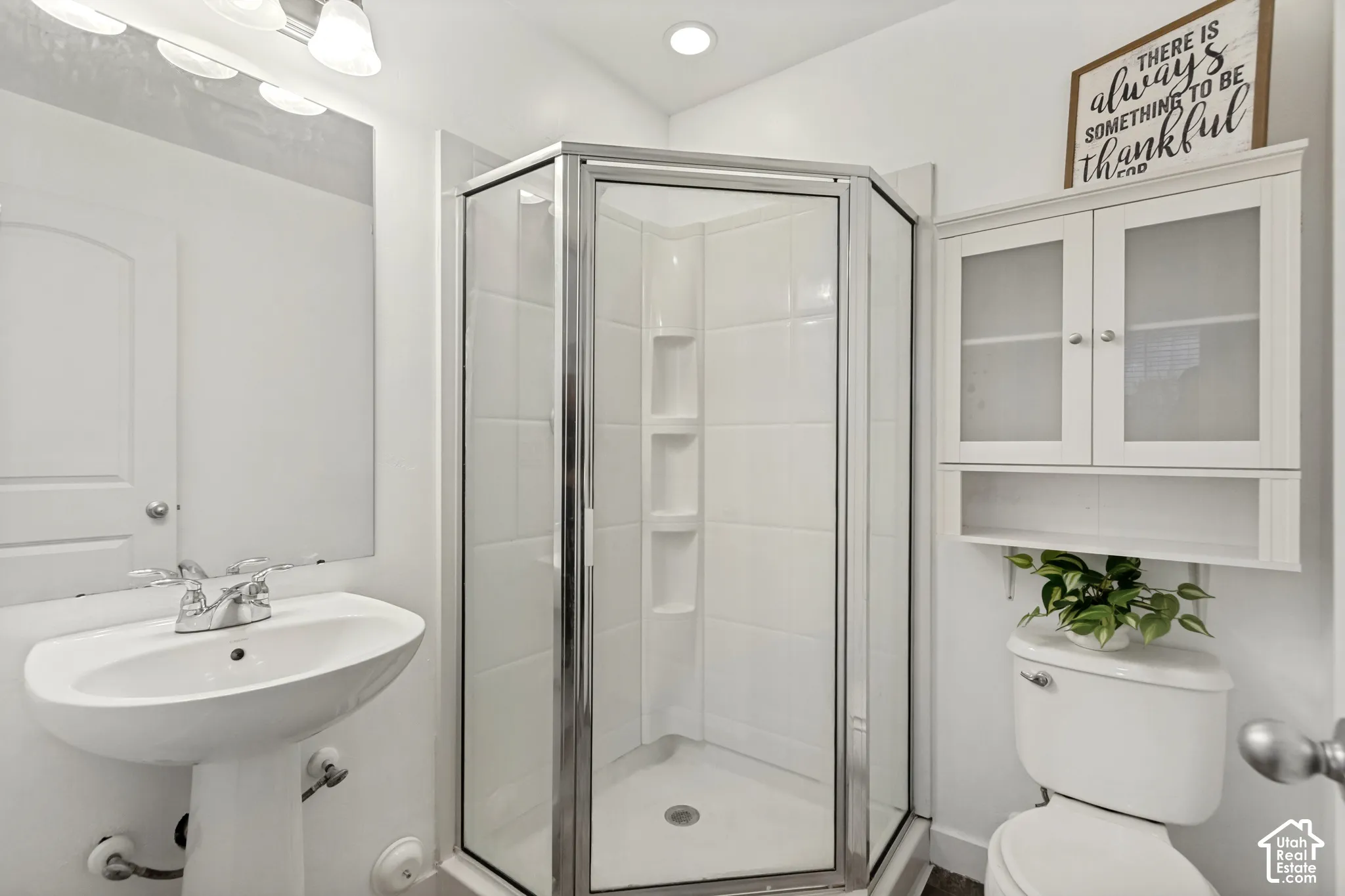 Bathroom featuring a corner shower with glass enclosure, pedestal sink with chrome faucet, wall-mounted cabinet with glass-front doors, and recessed lighting