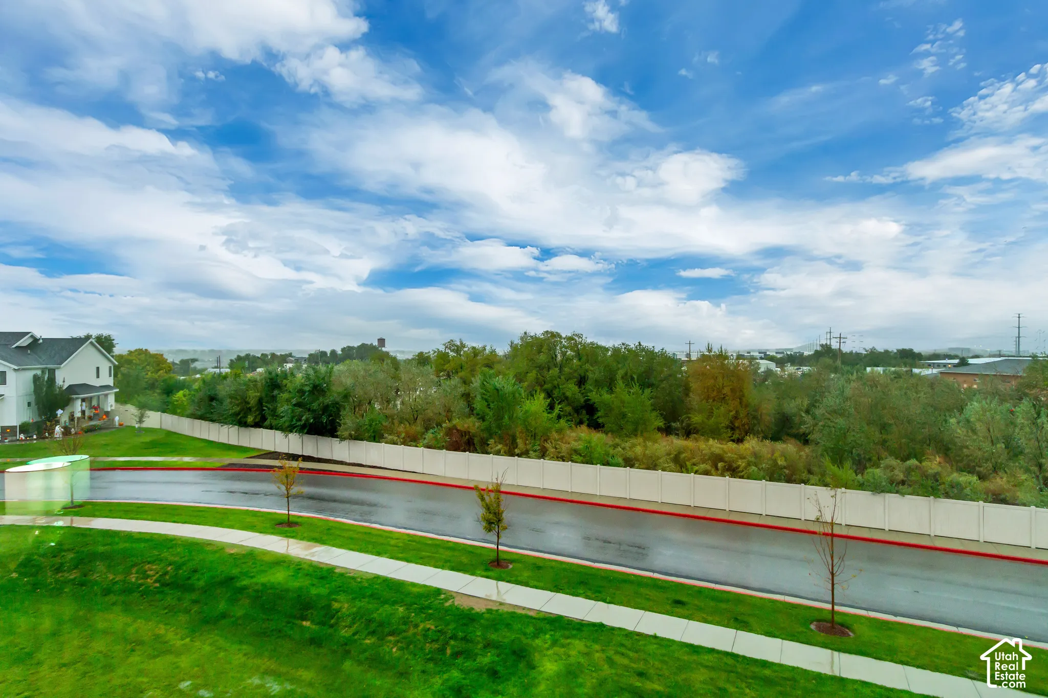 Paved roadway featuring concrete curbing, grass-lined sidewalk, perimeter fencing, and mature trees