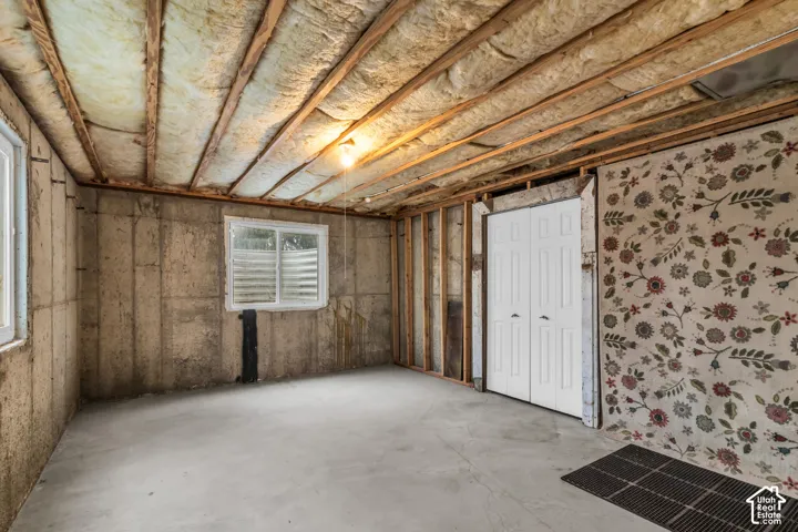 Unfinished utility space featuring exposed concrete foundation walls, ceiling joists with insulation, dual-pane windows, and a set of white bifold closet doors
