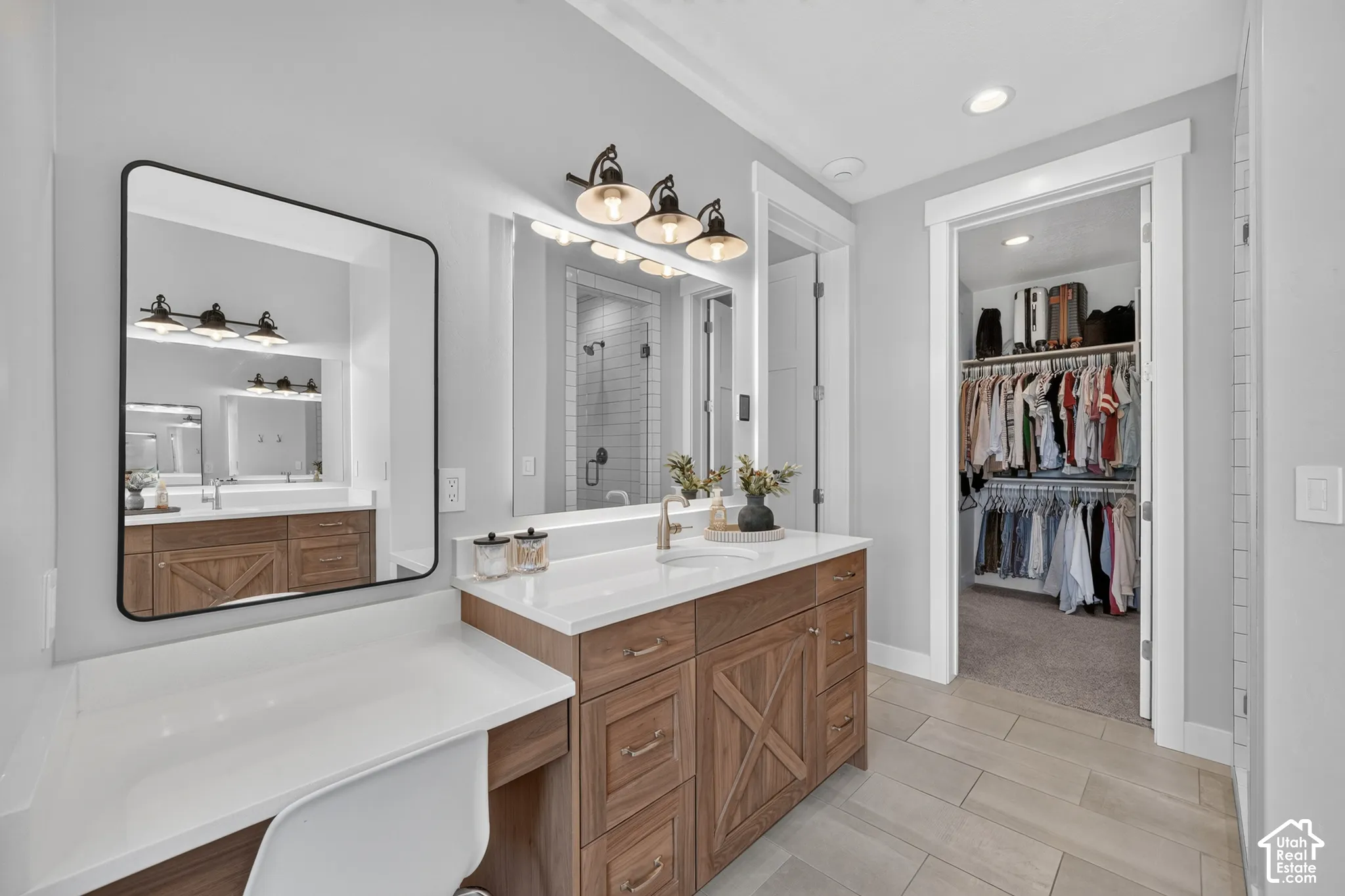 Wood-finish vanity featuring cross-brace detailing, white composite countertop with integrated sink, rectangular wall-mounted mirror, three-light metal fixture, and tile flooring