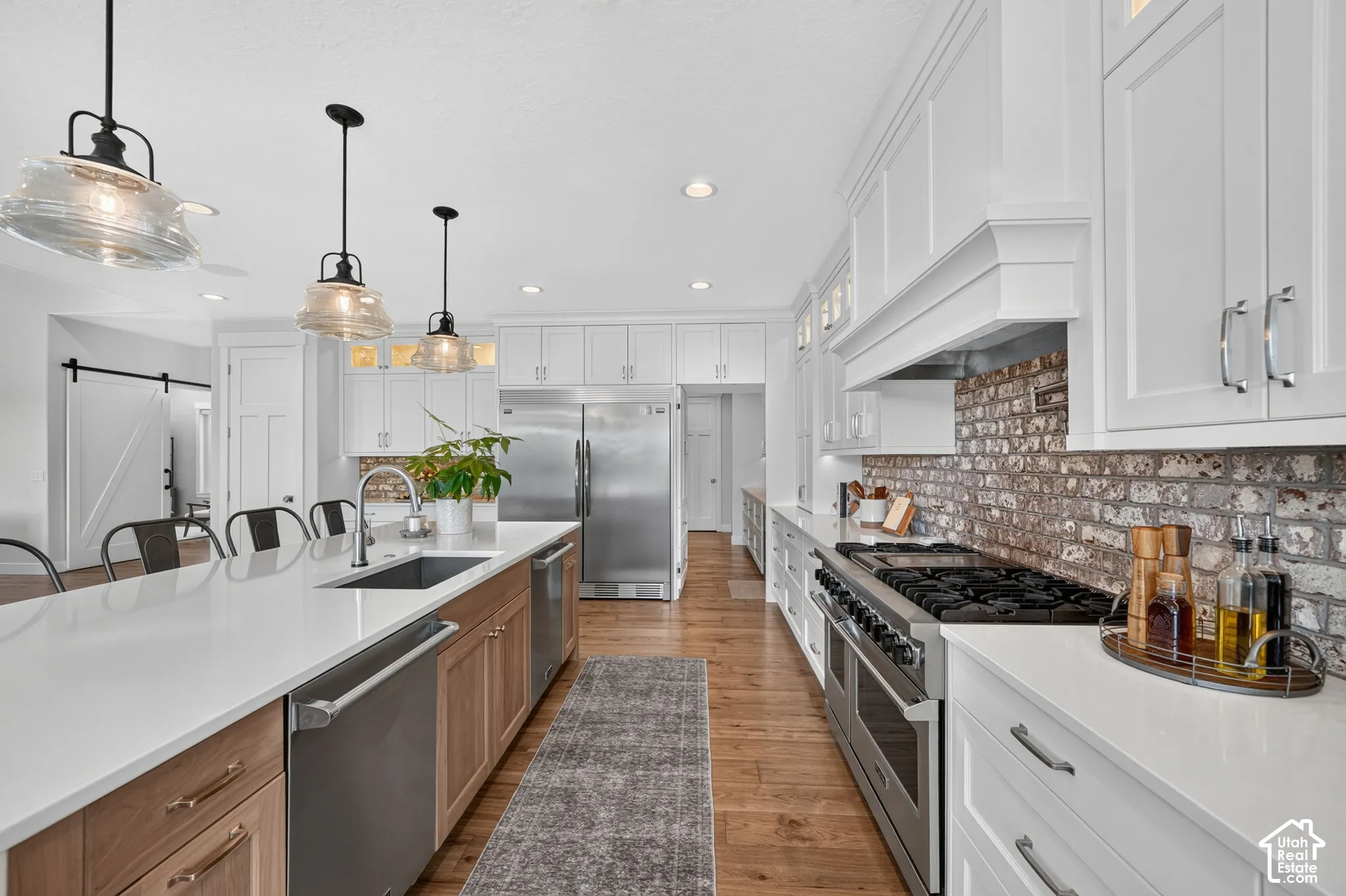Kitchen featuring a large central island with quartz countertops, custom white and wood-finish cabinetry, a professional-grade stainless steel range, exposed brick backsplash, and decorative pendant lighting