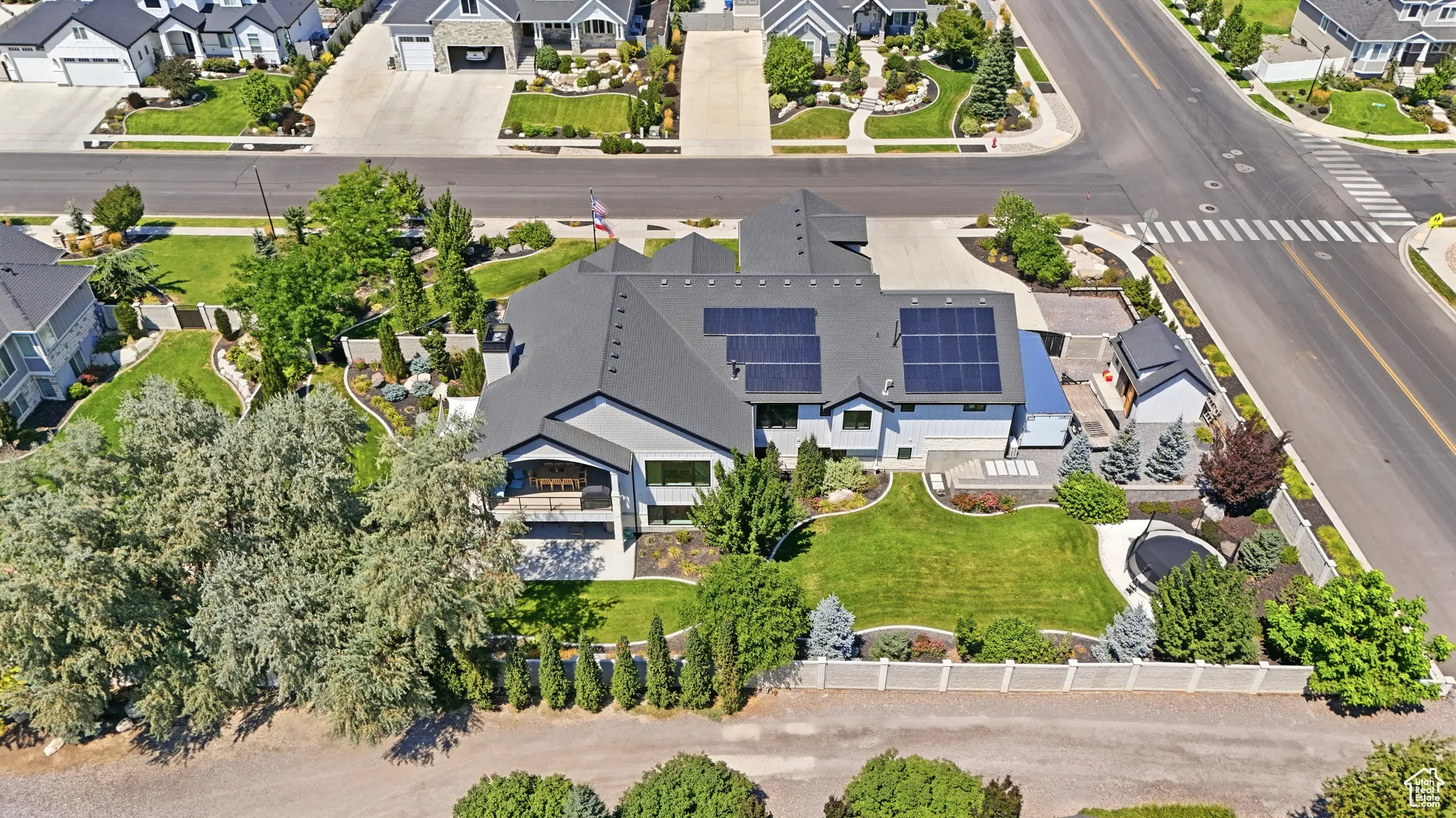 Aerial perspective of a multi-gabled residence featuring roof-mounted solar panels, a manicured lawn, perimeter fencing, and established trees