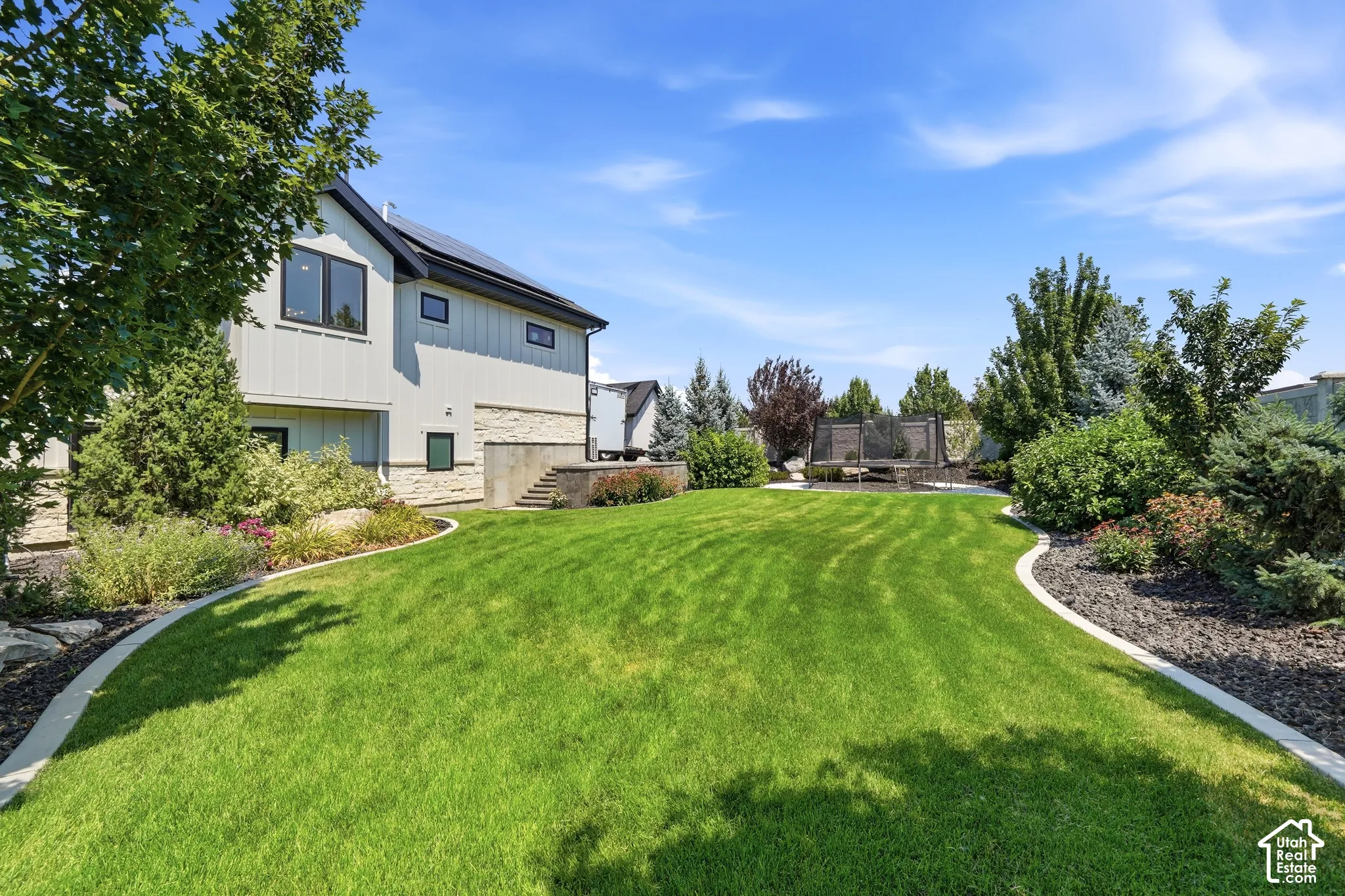 Expansive backyard featuring a manicured lawn, stone-accented exterior wall, concrete curbing, tiered stone stairs, and mature landscaping