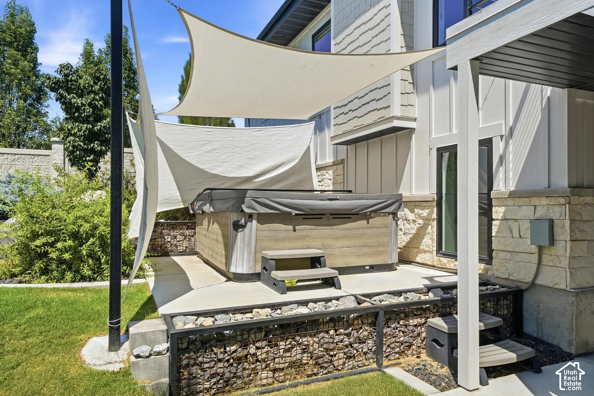 Outdoor living area featuring a built-in hot tub on a concrete patio, stone-accented exterior siding, and a metal-framed retaining wall filled with natural stone