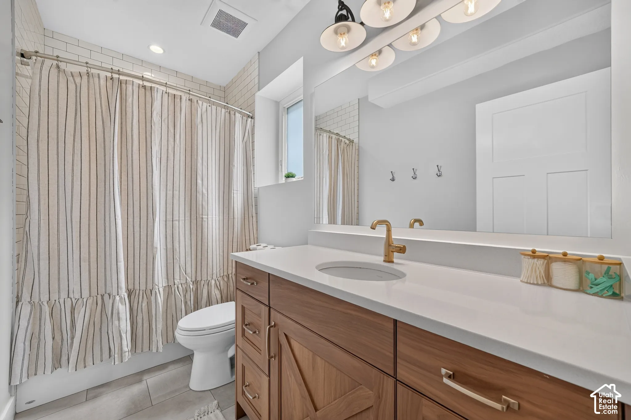Bathroom featuring a wood-finish vanity with a stone-composite countertop, undermount sink, brass-tone hardware, large-format tile flooring, and subway tile shower surround
