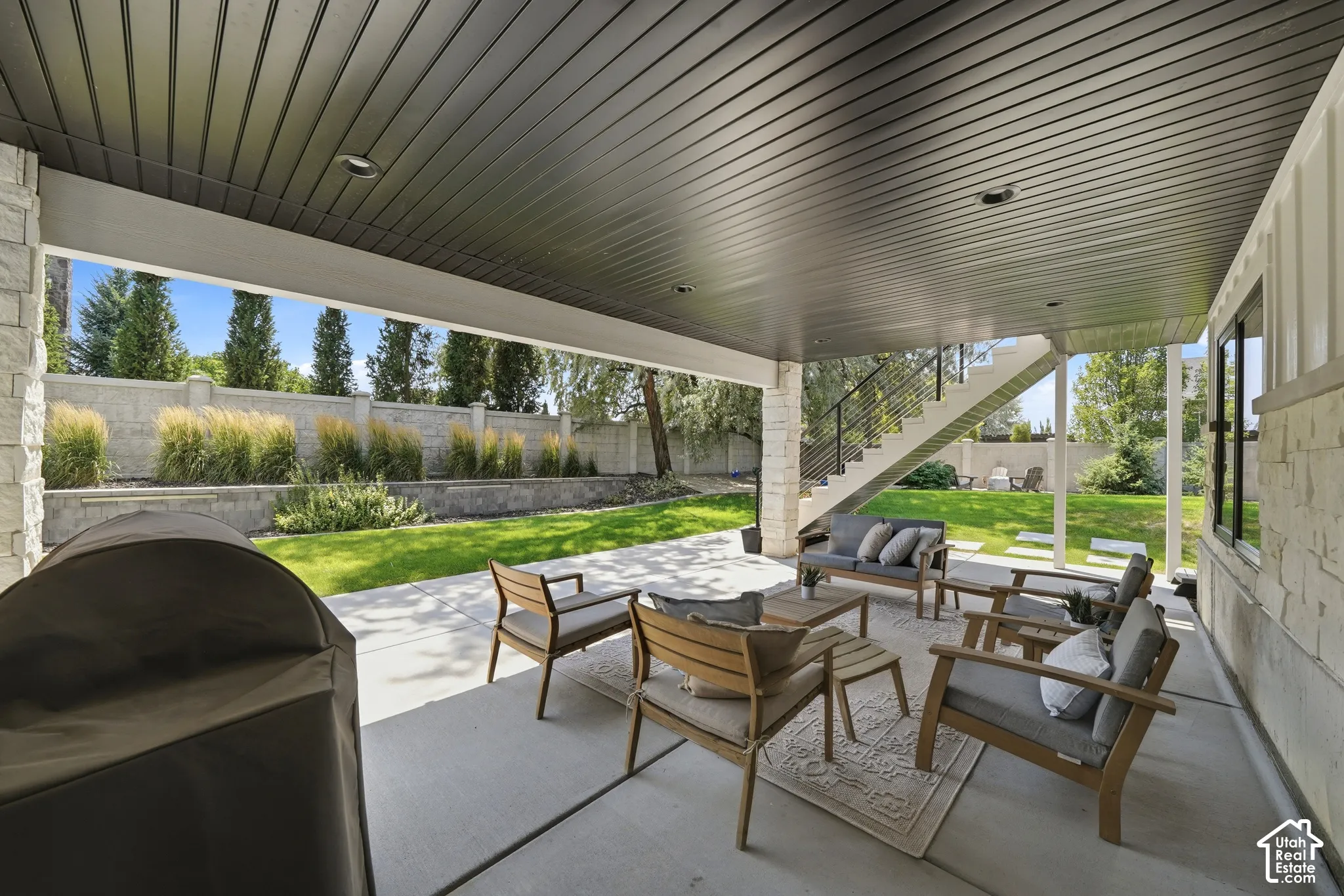 Covered concrete patio featuring recessed lighting, a stone-pillar support, built-in staircase with metal railings, and a terraced backyard with a perimeter wall