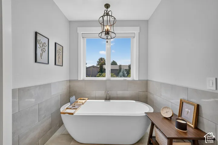 Freestanding soaking tub with a floor-mounted faucet, large-format tile wainscoting, polished metal lantern-style chandelier, and wide window casing