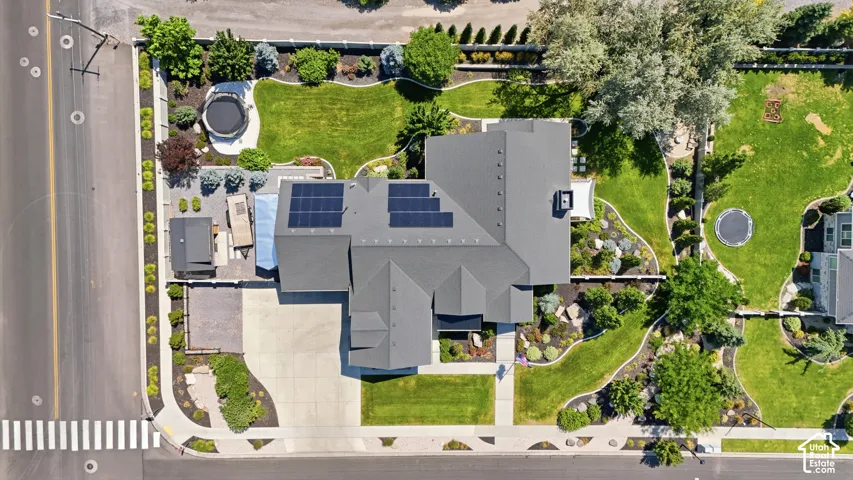 Aerial view featuring a residential property with roof-mounted solar panels, an attached multi-car garage, concrete driveway, and landscaped grounds with mature trees and perimeter fencing