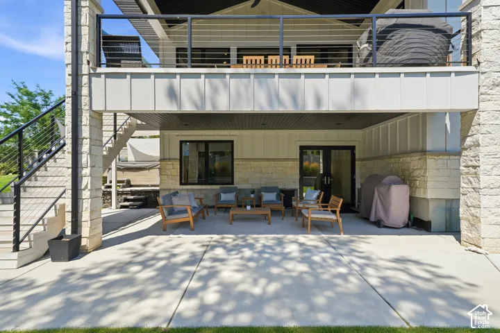 Covered ground-level patio with concrete flooring, stone-veneer accent walls, elevated balcony with cable railings, and exterior staircase