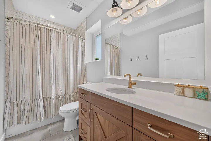 Bathroom featuring a wood-finish vanity with a stone-composite countertop, undermount sink, brass-tone hardware, large-format tile flooring, and subway tile shower surround