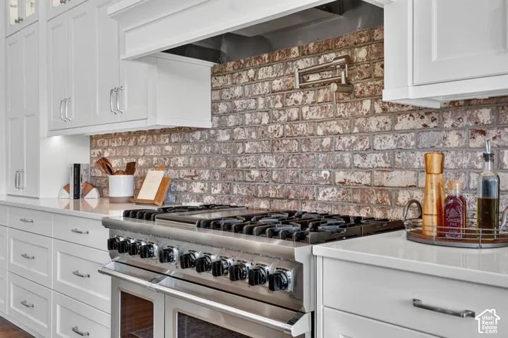 White shaker-style cabinetry, professional-grade stainless steel gas range, textured brick backsplash, wall-mounted pot filler, and solid-surface countertops