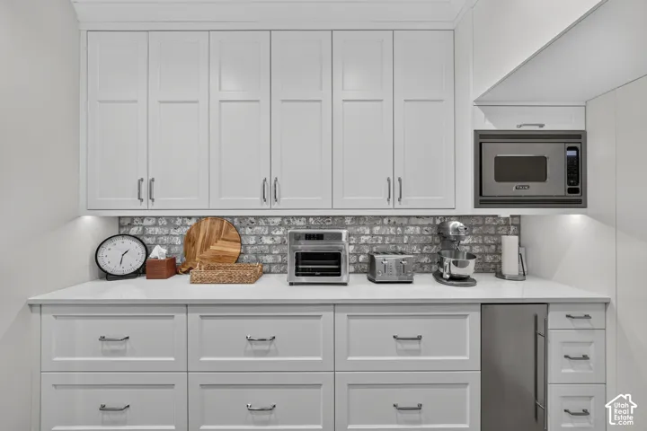 Custom white shaker-style cabinetry with satin nickel hardware, solid-surface white countertops, textured brick backsplash, and built-in stainless steel microwave