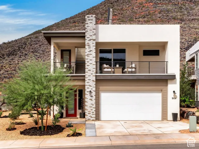 Modern two-story residence featuring a stucco exterior, stone-veneer chimney, covered second-story balcony with metal railings, and desert-inspired xeriscaped landscaping against a mountain backdrop