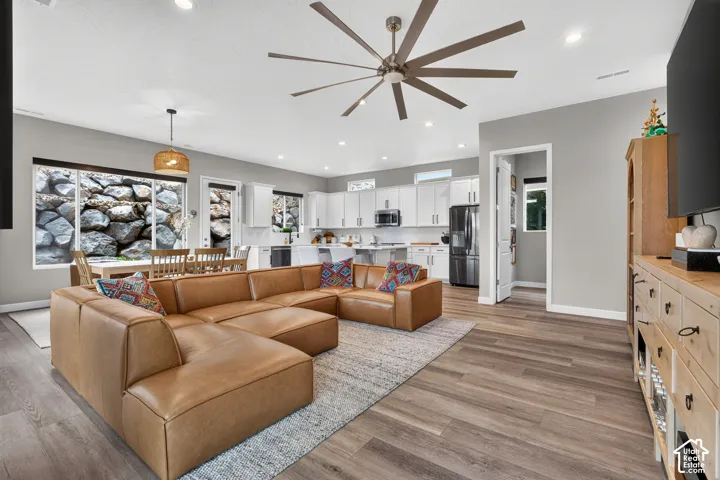 Open-concept interior featuring wood-finish flooring, white cabinetry, a multi-blade ceiling fan, recessed lighting, and large windows with natural rockery visibility