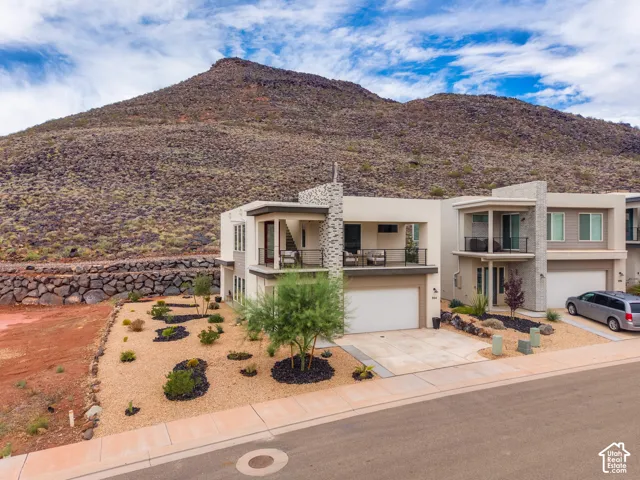 Contemporary two-story exterior featuring stucco siding, stone veneer accents, a second-story balcony with metal railing, and a concrete driveway set against a mountain backdrop