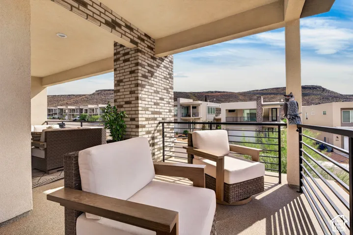 Covered balcony featuring a textured concrete floor, brick-accented pillar, recessed lighting, and metal horizontal railing, set against a mountainous backdrop